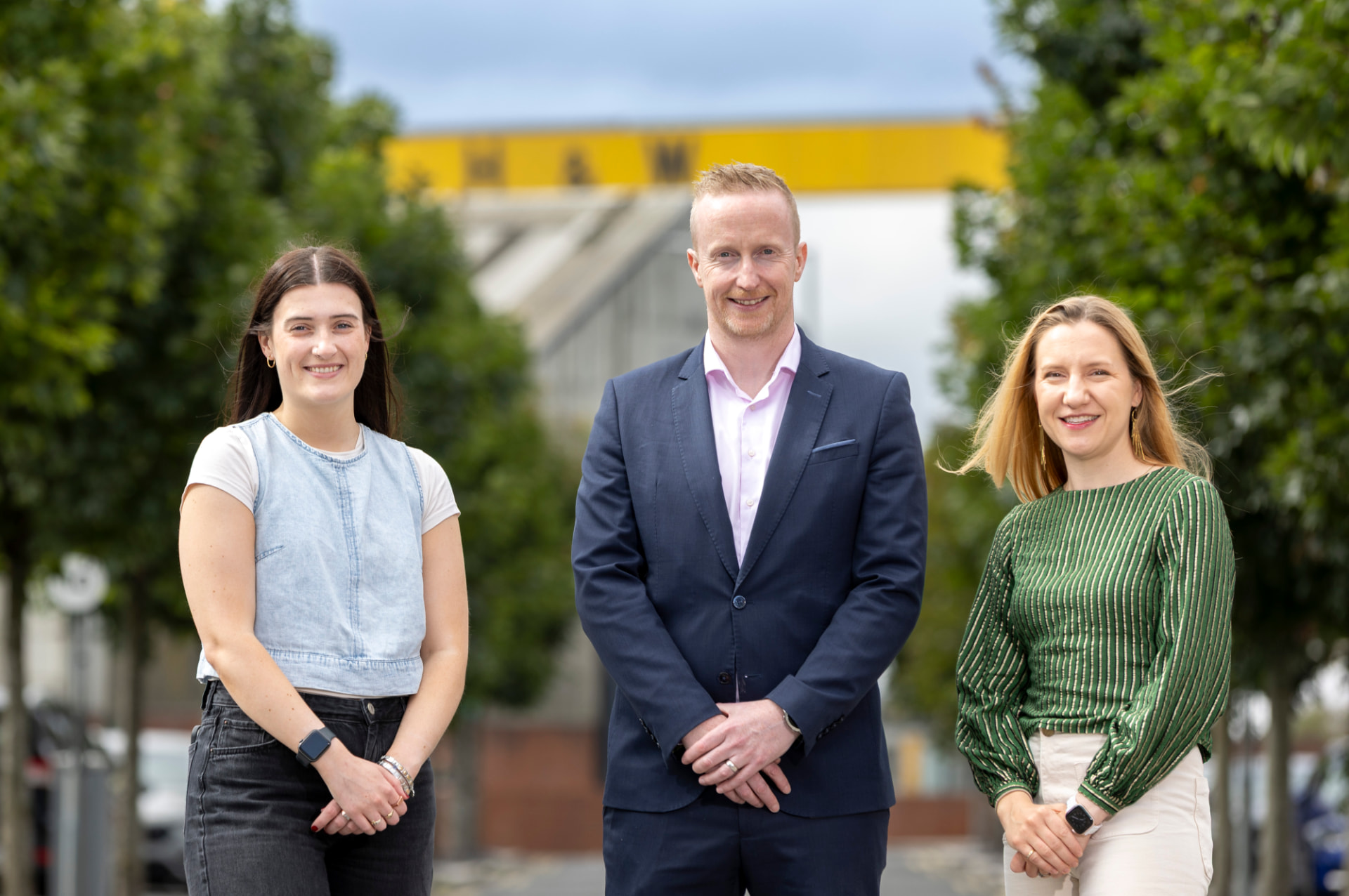 Three members of the SEAM team standing on the Newtownards Road in east Belfast, with the Harland & Wolff crane behind them